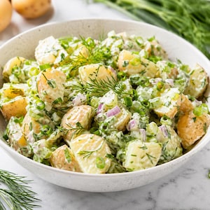 May include: A bowl of potato salad with dill, green onions, and red onion. The potatoes are cut into bite-sized pieces and mixed with a creamy dressing. Fresh dill sprigs and sliced red onion are arranged around the bowl.