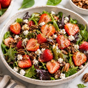 Op de afbeelding: Een frisse salade met gesneden aardbeien, pecannoten en verkruimelde kaas in een witte kom. De salade staat op een wit oppervlak, met extra aardbeien in een aparte kom.