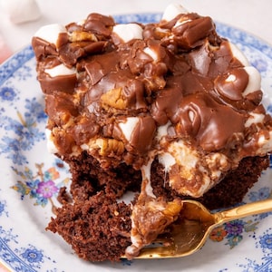 May include: A square slice of chocolate cake topped with melted chocolate, marshmallows, and pecans, served on a floral patterned plate. A gold fork is partially inserted into the cake, with a bite taken out.