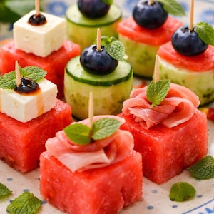 Puede incluir: Un plato de coloridos aperitivos con cubos de sandía, pepino y queso. Cada cubo está cubierto con varios ingredientes como arándanos, prosciutto o glaseado balsámico, y adornado con hojas de menta. Brochetas de madera sujetan los ingredientes.