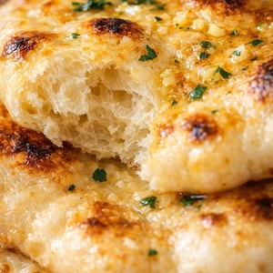 Peut inclure: Gros plan sur du pain naan doré et fraîchement cuit, avec une bouchée. Le pain est garni d'ail et d'herbes, avec un intérieur moelleux et aéré. L'image met en évidence la texture et les détails du pain.