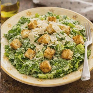 Puede incluir: Una ensalada César en un cuenco decorativo. La ensalada contiene lechuga romana, picatostes y queso parmesano rallado. Un tenedor descansa en el lateral del cuenco. Una botella de aceite es visible en el fondo.