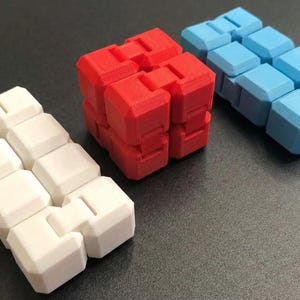 May include: Three colourful fidget toys in white, red, and blue. The toys are made of connected, geometric blocks that can be manipulated. The red toy is assembled into a cube, while the white and blue toys are in a linear configuration.