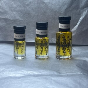 May include: Three glass perfume bottles with black caps and labels. The bottles contain a yellow liquid. The labels read "Damask Oil" and "Isk of Isfahan". The bottles are arranged in a row on a textured surface.
