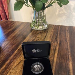 Puede incluir: Una moneda de plata en un estuche de presentación negro. La moneda presenta texto y un diseño, y el estuche está etiquetado como "The Royal Mint". Un ramo de rosas en un jarrón de cristal se encuentra en el fondo sobre una superficie de madera.