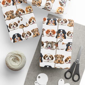 Könnte beinhalten: Geschenkpapier mit einem sich wiederholenden Muster von Welpengesichtern im Aquarellstil in verschiedenen Rassen und Farben. Die verpackten Geschenke sind mit Bindfaden und leeren Anhängern gebunden. Ein Knäuel Bindfaden, eine Schere und Anhänger sind ebenfalls sichtbar.