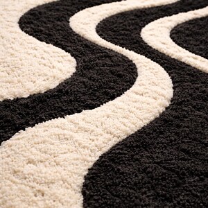May include: A close-up of a rug with a wavy pattern. The rug features alternating bands of black and cream colors. The texture appears soft and plush, with a high pile. The design is a repeating, flowing pattern.