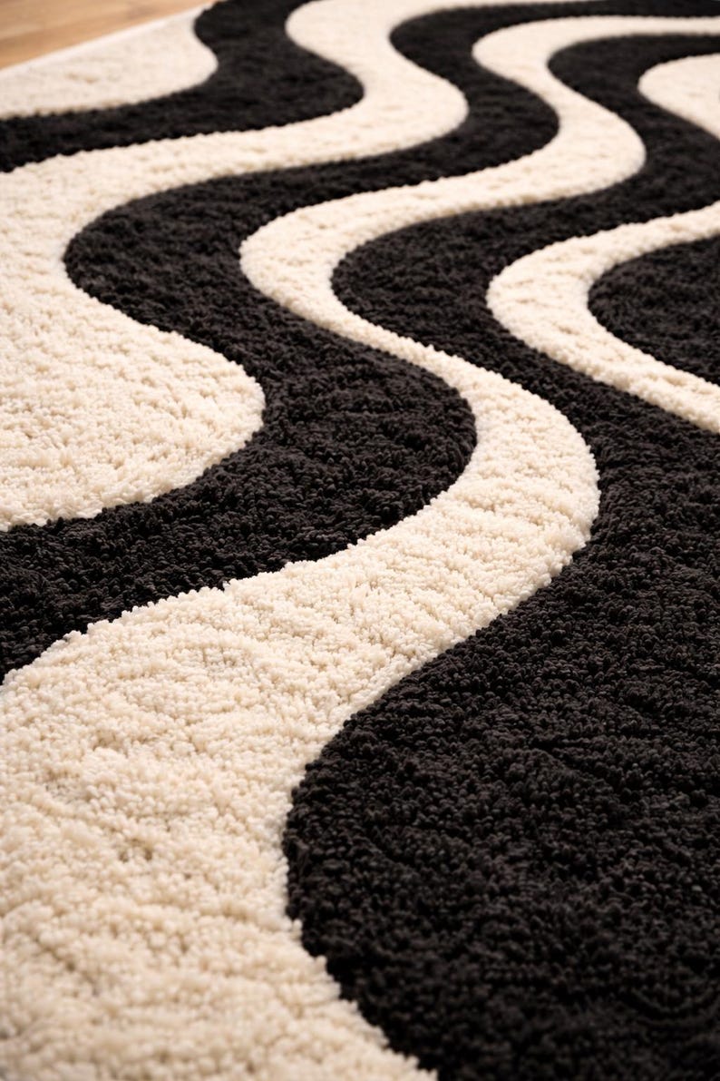 May include: A close-up of a rug with a wavy pattern. The rug features alternating bands of black and cream colors. The texture appears soft and plush, with a high pile. The design is a repeating, flowing pattern.