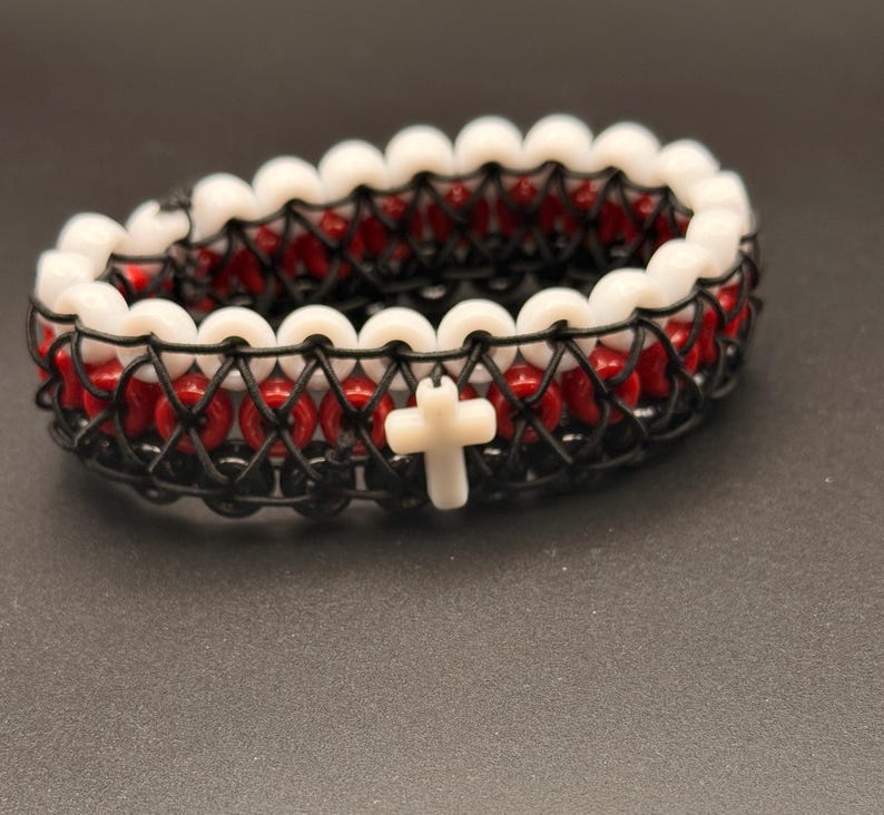 Puede incluir: Una pulsera de cuentas con cuentas rojas y blancas, con un dise&ntilde;o tejido en negro. La pulsera tiene un dije de cruz blanca. La pulsera est&aacute; sobre una superficie gris oscuro.