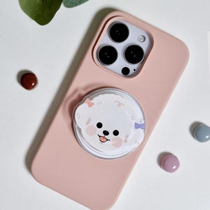 May include: A light pink phone case with a cartoon dog pop socket. The pop socket features a white dog with pink and blue bows. The phone case has a triple-lens camera and is surrounded by small, colorful, round objects.