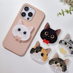 May include: A pink phone case with a white cat face phone grip attached. Four other cat face phone grips are visible, each with a different pattern and colour. Each cat face has a bow tie.