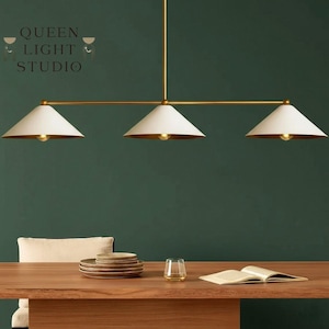 May include: A three-light pendant lamp with a gold finish and white cone-shaped shades. The lamp is suspended from the ceiling and hangs over a wooden table. The text "QUEEN LIGHT STUDIO" is visible in the upper left corner.