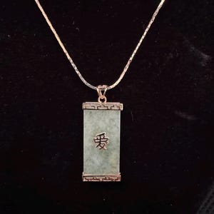 May include: A silver necklace with a rectangular jade pendant. The pendant has the Chinese character for "love" engraved on it. The jade is a light green color, and the pendant is framed in a silver setting. The necklace is on a black background.