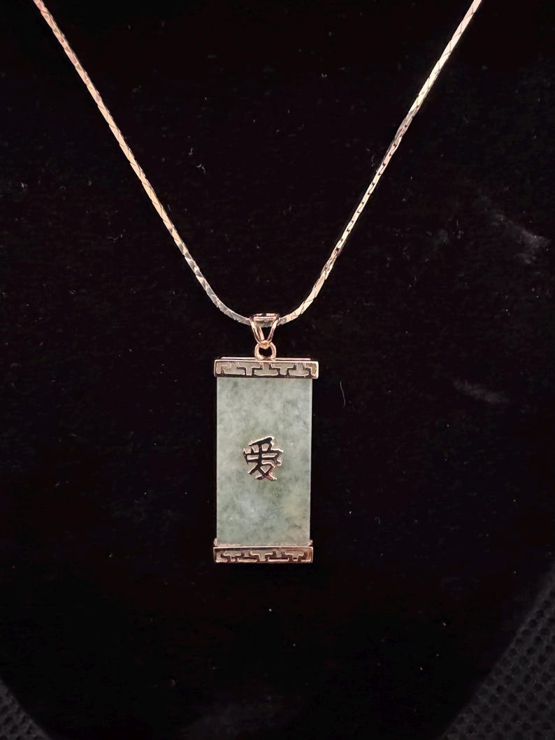 May include: A silver necklace with a rectangular jade pendant. The pendant has the Chinese character for "love" engraved on it. The jade is a light green color, and the pendant is framed in a silver setting. The necklace is on a black background.