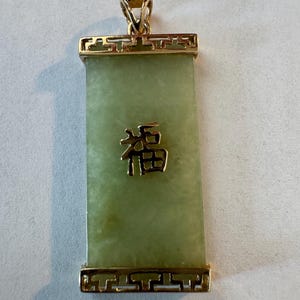 May include: Rectangular jade pendant with a gold-colored frame and bail. The pendant has a light green hue with a gold-colored Chinese character in the center. The frame features a decorative design.