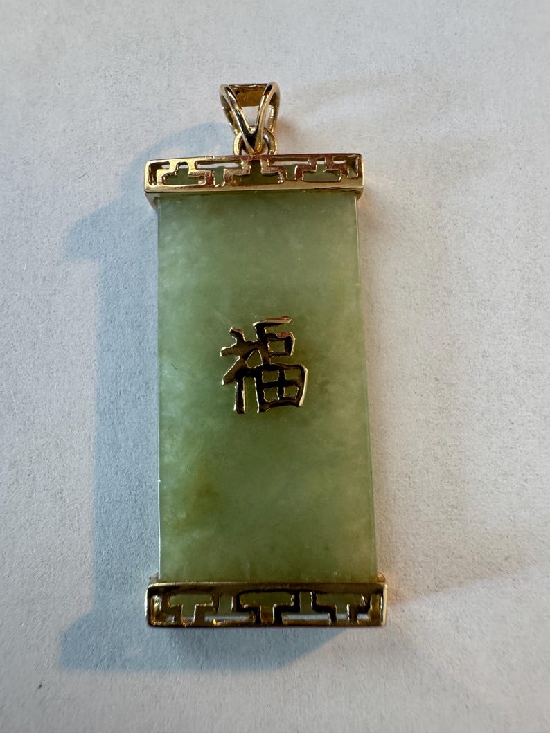 May include: Rectangular jade pendant with a gold-colored frame and bail. The pendant has a light green hue with a gold-colored Chinese character in the center. The frame features a decorative design.