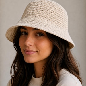 May include: A cream-colored bucket hat featuring a dense, uniform crochet stitch pattern. The hat has a rounded crown and a downward-sloping brim that encircles the head, constructed from a single continuous yarn in a neutral, off-white shade.