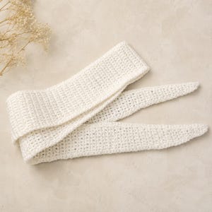 Puede incluir: Una diadema o accesorio para el cabello largo de ganchillo en color blanco roto con un patrón de tejido texturizado. El accesorio tiene una sección central ancha y rectangular que se estrecha en dos extremos largos y puntiagudos.