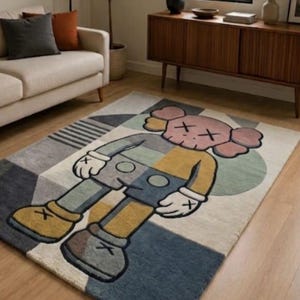 May include: A rectangular rug with a cartoon character design. The rug has a geometric pattern in grey, blue, yellow, and cream. The cartoon character is in the centre, with pink, yellow, and blue colours.