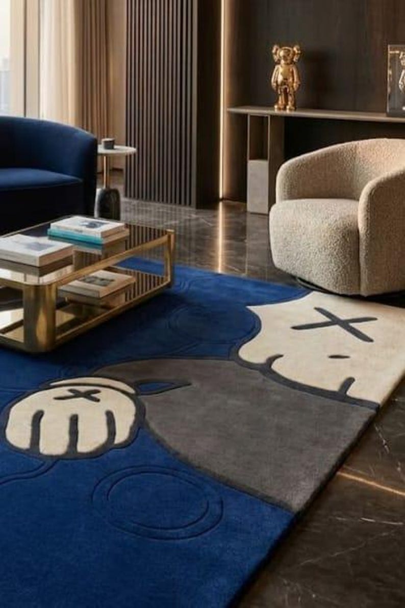 Peut inclure: Tapis moderne avec un motif de personnage de dessin anim&eacute; en bleu, gris et cr&egrave;me. Le tapis est dans un salon avec une table basse dor&eacute;e, un canap&eacute; bleu et un fauteuil beige. Le design du tapis est une caract&eacute;ristique cl&eacute;.