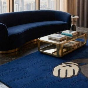 Peut inclure: Salon moderne avec un canap&eacute; incurv&eacute; bleu marine, une table basse en verre avec cadre dor&eacute; et un tapis bleu avec un motif de personnage de dessin anim&eacute;. La pi&egrave;ce a de grandes fen&ecirc;tres avec vue sur la ville et des rideaux de couleur neutre.
