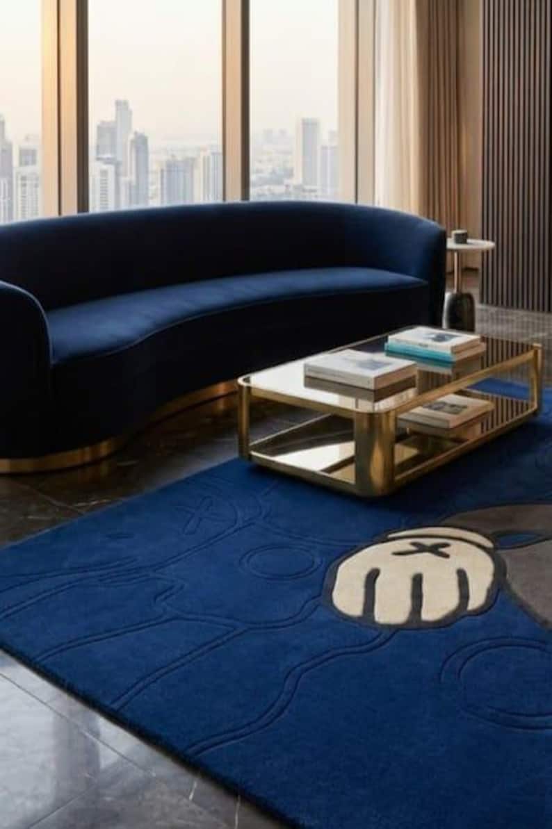 Peut inclure: Salon moderne avec un canap&eacute; incurv&eacute; bleu marine, une table basse en verre avec cadre dor&eacute; et un tapis bleu avec un motif de personnage de dessin anim&eacute;. La pi&egrave;ce a de grandes fen&ecirc;tres avec vue sur la ville et des rideaux de couleur neutre.