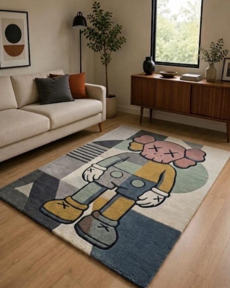 May include: A rectangular rug with a cartoon character design in grey, blue, yellow, and pink. The rug is placed on a wooden floor in a living room, with a sofa and artwork in the background. The rug's design is modern and geometric.