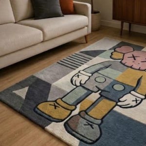 May include: Rectangular rug with a cartoon character design in blue, grey, yellow, and pink. The rug has a geometric pattern and is placed on a wooden floor. The rug's dimensions are in centimetres.