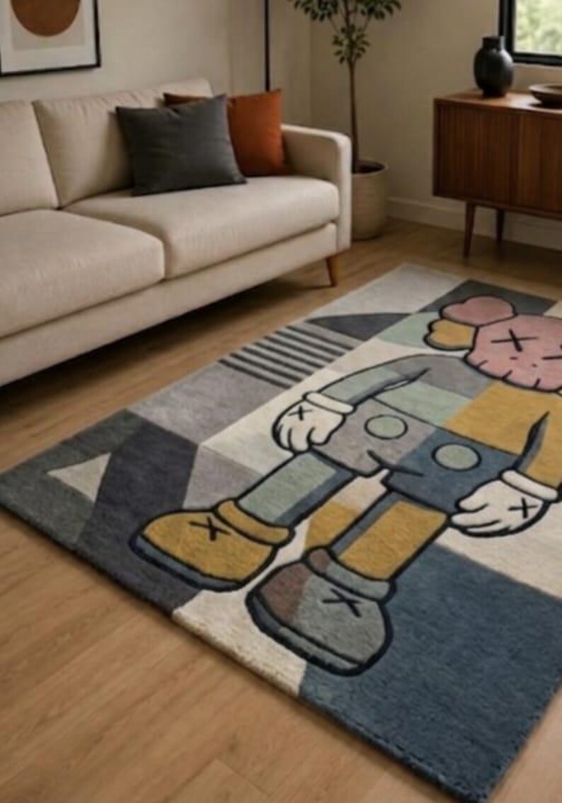 May include: Rectangular rug with a cartoon character design in blue, grey, yellow, and pink. The rug has a geometric pattern and is placed on a wooden floor. The rug's dimensions are in centimetres.