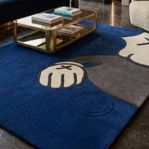 Peut inclure: Un tapis bleu, gris et cr&egrave;me avec un motif de personnage de dessin anim&eacute;. Le tapis pr&eacute;sente une grande main avec un X au dos. Le tapis est pos&eacute; sur un sol en marbre fonc&eacute;, avec une table basse dor&eacute;e et un canap&eacute; bleu en arri&egrave;re-plan.