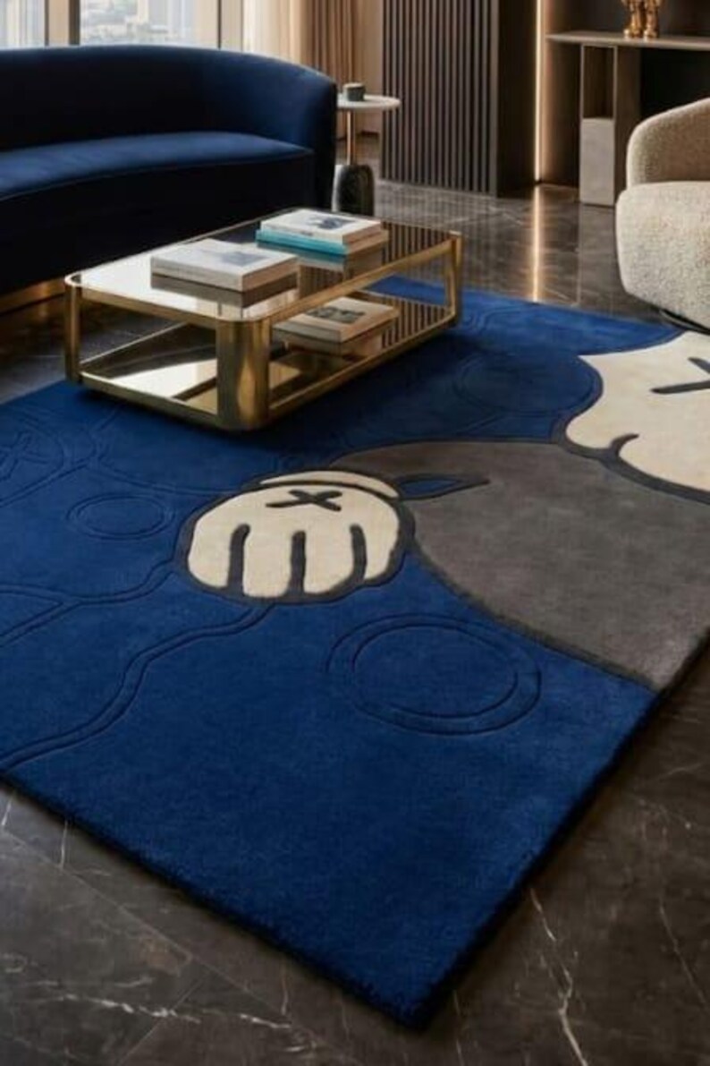 Peut inclure: Un tapis bleu, gris et cr&egrave;me avec un motif de personnage de dessin anim&eacute;. Le tapis pr&eacute;sente une grande main avec un X au dos. Le tapis est pos&eacute; sur un sol en marbre fonc&eacute;, avec une table basse dor&eacute;e et un canap&eacute; bleu en arri&egrave;re-plan.