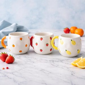 Könnte beinhalten: Drei weiße Keramikbecher mit Fruchtdesign. Ein Becher hat orangefarbene Griffe und Orangendesigns, ein anderer hat rote Griffe und Erdbeerdesigns, und der dritte hat gelbe Griffe und Zitronendesigns.