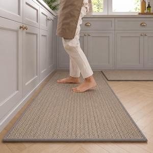 Op de afbeelding: Een rechthoekig, beige keukenkleed met een grijze rand. Het kleed heeft een geribbelde textuur en ligt op een houten vloer. Een kleiner bijpassend kleed is zichtbaar op de achtergrond. Een persoon staat op het kleed.