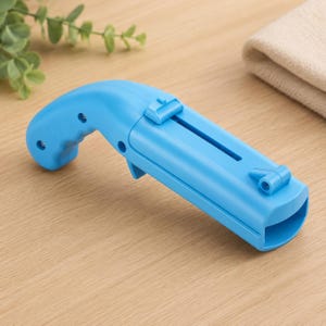 May include: A light blue plastic tool with a handle and a sliding mechanism. The tool has a trigger-like design and a slot running along its length. It is resting on a wooden surface.