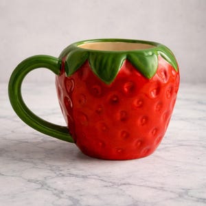 Könnte beinhalten: Eine keramische Tasse in Erdbeerform. Die Tasse ist rot mit einem grünen Henkel und Oberteil sowie einem hellbeigen Innenraum. Der rote Körper hat kleine, strukturierte Unebenheiten. Sie steht auf einer Marmoroberfläche.
