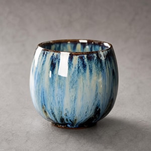 Könnte beinhalten: Eine Keramiktasse mit glänzendem Finish. Die Tasse hat eine Mischung aus Blau- und Weißtönen, mit dunkleren blauen Streifen und einem braunen Rand. Die Tasse hat eine abgerundete Form und einen kleinen Sockel.