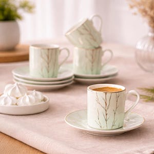 Könnte beinhalten: Ein Set kleiner, mintgrüner Kaffeetassen und Untertassen mit goldenem Rand und einem Baumzweig-Design. Die Tassen sind mit Kaffee gefüllt. Ein Teller mit weißen Baisers befindet sich im Vordergrund.