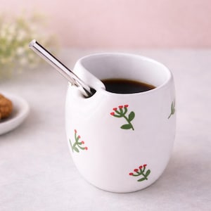 Könnte beinhalten: Weißer Keramikbecher mit einzigartigem Design, gefüllt mit dunkler Flüssigkeit und einem silbernen Strohhalm. Der Becher ist mit grünen Blättern und roten Blumenillustrationen verziert. Ein kleiner Teller mit einem Gebäckstück befindet sich im Hintergrund.