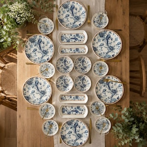 Può includere: Un set di stoviglie con un motivo chinoiserie blu e bianco raffigurante uccelli e motivi floreali. La collezione comprende piatti piani rotondi, piatti da insalata, ciotole piccole e piatti da portata rettangolari disposti su un tavolo.