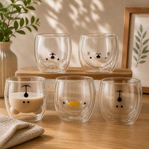 Könnte beinhalten: Fünf klare doppelwandige Gläser mit Cartoon-Tiergesichtern. Ein Glas enthält eine hellbraune Flüssigkeit. Die Gläser stehen auf einer Holzoberfläche, mit einer Vase mit Grünzeug und einem gerahmten Bild im Hintergrund.