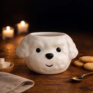 Könnte beinhalten: Eine weiße Keramik-Tasse in Form eines Hundekopfes mit schwarzen Augen und Nase sowie einem lächelnden Mund. Die Tasse steht auf einer Holzoberfläche, im Hintergrund befinden sich ein goldener Löffel, Kekse und Kerzen. Ein dekoratives Objekt.