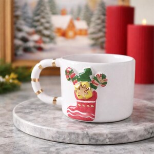 Könnte beinhalten: Weiße Keramiktasse mit einem Weihnachtsstrumpf-Design. Die Tasse zeigt einen rot-grünen Strumpf mit einem festlichen Elf. Der Griff ist weiß mit goldenen Streifen. Zwei rote Kerzen sind im Hintergrund.