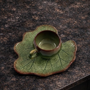 Könnte beinhalten: Ein Keramik-Teebecher- und Untertassen-Set. Die Tasse und die Untertasse sind grün mit einem Blattmuster. Die Tasse hat einen braunen Rand und Henkel. Die Untertasse ist wie ein Blatt geformt. Das Set steht auf einer dunkelbraunen Oberfläche.