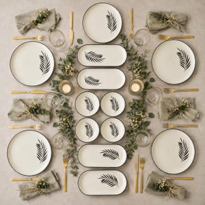 Könnte beinhalten: Ein Geschirrset mit cremefarbenen Tellern, ovalen Platten und kleinen Schalen, die jeweils mit einem schwarzen Farnblattmotiv verziert sind. Das Set enthält goldfarbene Gabeln und Messer, klare Gläser sowie mit Grün gebundene Leinenservietten.