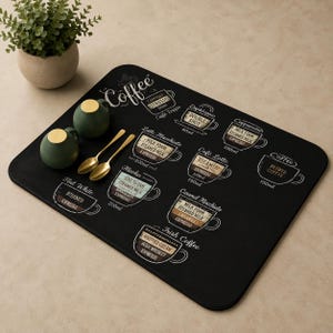 Puede incluir: Un mantel individual rectangular negro con ilustraciones de bebidas de café en blanco. Dos tazas de cerámica verde con tapas doradas y dos cucharas doradas están sobre la alfombrilla. Una pequeña planta en maceta está en el fondo.