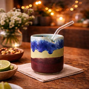 Könnte beinhalten: Ein handgefertigter Keramikbecher mit einem Glasstrohhalm. Der Becher hat ein mehrschichtiges Design mit blauen, gelben und burgunderfarbenen Farben. Der Becher steht auf einer beigen Leinen-Serviette auf einem Holztisch.