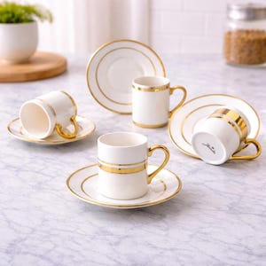 Könnte beinhalten: Weiße Kaffeetassen und Untertassen aus Keramik mit goldenen Verzierungen. Die Tassen haben eine zylindrische Form mit einem goldenen Henkel und einem goldenen Band in der Mitte. Die Untertassen haben einen goldenen Rand. Das Set wird auf einer Marmoroberfläche präsentiert.