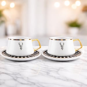 Könnte beinhalten: Zwei weiße Keramik-Kaffeetassen mit goldenen Griffen und Rändern, jede mit einem schwarzen Hirschkopf-Design. Sie stehen auf passenden Untertassen mit schwarz-weiß gestreiften Mustern. Das Set steht auf einer Marmoroberfläche.
