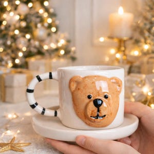 Könnte beinhalten: Eine weiße Keramiktasse mit einem 3D-Bärengesicht-Design, einer passenden Untertasse und einem Griff mit schwarzen Streifen. Die Tasse wird vor einem verschwommenen Hintergrund mit Weihnachtsbeleuchtung und einer Kerze gehalten.