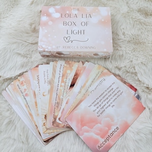 Puede incluir: Un juego de cartas de oráculo con una caja. La caja es de color rosa claro con el texto "THE LOLA LIA BOX OF LIGHT" y "BY REBECCA DOWNING". Las cartas son de color rosa claro y blanco con texto inspirador. La palabra "Acceptance" es visible en una carta.